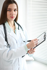 Young female doctor filling up medication history recors while standing near the window in hospital