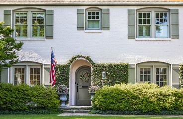 Vine covered entrance to white painted brick house with arched front door and wreath and arched windows with green shutters - landscaped with American flag