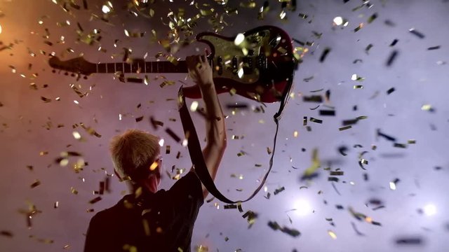 GUITARIST ON SCENE raises the guitar up, spectators applaud. The guitarist performs on stage. Stage light, smoke. From above fall golden confetti, Slow motion instrument playing band of men.