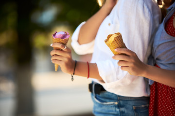 Close up shot of ice cream cones in hand of a woman standing with her friend. Two young women outdoors eating icecream on a sunny day. Isolated view, no face, copyspace for designers. Summer theme.