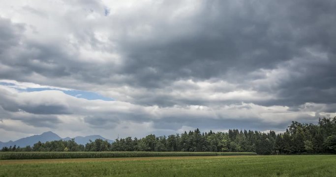 Time lapse of a grass and corn field with forest in the back. Darker clouds can be seen moving above with occasional sun breaking through. Alps in the background.