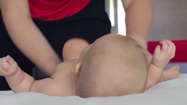 A six months old baby girl is lying on white sheets close to her mother, who is standing on the knees on the same bed. The woman is gently rolling baby from side to side, doing gymnastics with her