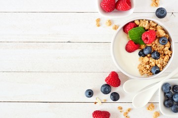 Healthy yogurt with raspberries, blueberries and granola. Above view, side border over a white wood background. Copy space.