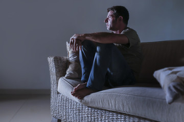 lifestyle dramatic light portrait of young sad and depressed man sitting at shady home couch in pain and depression feeling lost looking away thoughtful