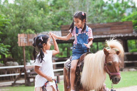 Cute asian child girl riding a pony and making hi five gesture with her elder sister in the farm with fun