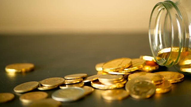 Yellow coins fell out from jar. Symbol of investing, keeping money concept. Collecting cash conis in glass tin as moneybox. Close-up still life with gold coins on black table and rotating penny.