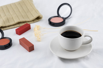 A cup of coffee with cosmetics on white bed, selective focus on coffee