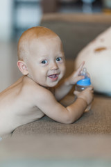 A child of 8 months is at the sofa with a bottle of water, smiling in a diaper
