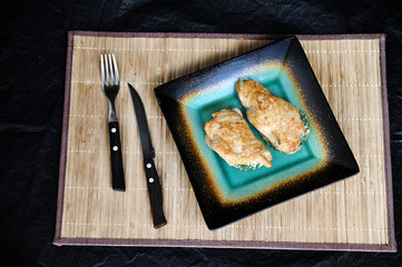 Chicken breast stuffed with ricotta and spinach. Next to the plate are cutlery. Under the plate is a bamboo spfetka. Dark background. View from above. 
