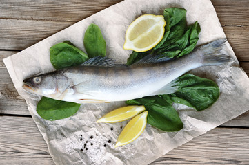 Raw pikeperch on parchment paper. Next to the fish are fresh spinach and lemon slices. Light wooden background. View from above. Close-up.