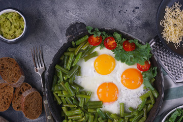 Fried eggs in an old pan with tomatoes, avocado pesto, green beans, pepper and salad on a dark background