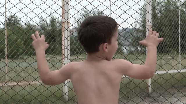 The boy with no shirt on shaking the mesh fence. Muscular healthy body. Summer beach. Restriction of freedom. Children's game. Portrait from the waist up. One person. The trees in the background.