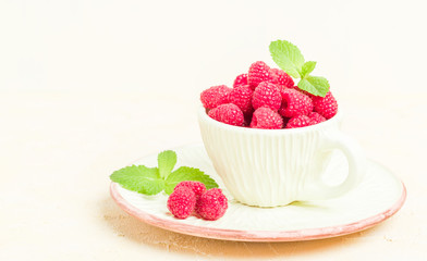 Ripe raspberries with green mint leaves in cup and saucer on pastel yellow background.