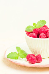 Ripe raspberries with green mint leaves in cup and saucer on pastel yellow background.