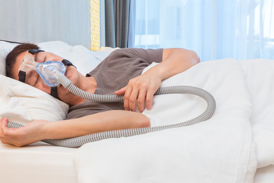 Asian man wearing CPAP mask, connecting to air hose, sleeping on his bed in his bedroom