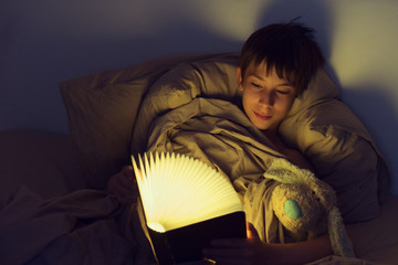 Adorable boy reads book before sleeping in bed at home. Happy childhood.
