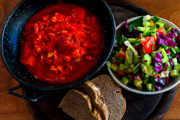 Shakshuka with eggs, tomato, and carrots in iron pan, vegetables salad and fresh bread. Shakshuka - traditional Israeli tomato stew with eggs.Breakfast Israeli in restaurant.