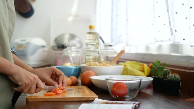 4K footage. woman's hand slice tomato with recipe book open at foreground, prepare ingredients for cooking at kitchen counter.
