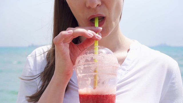 Close up of vegan woman in white shirt drinking strawberry smoothie from plastic cup against sea in slow motion. Fit vegetarian female enjoying healthy lifestyle outdoors