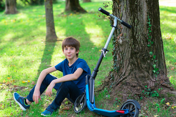 Boy sitting and resting near tree with his scooter, summer nature outdoor. image toned and noise...