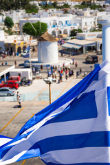 Fototapeta na wymiar Greek flag over Paros harbor with wind mill, Cyclades, Greece. Theme of Cyclades, Greece.