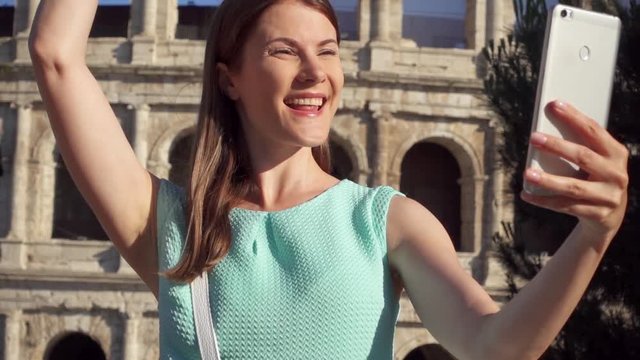 Woman do selfie on mobile phone near famous attraction Colosseum in Rome, Italy. Teenage girl smiling in slow motion. Happy female tourist enjoying her european vacation. Student travel through Europe