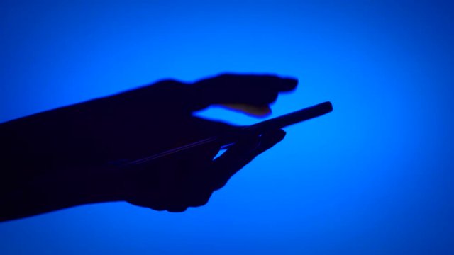 Close-up of silhouette of woman hands using smartphone touchscreen on blue background. Black contour of palms shadow typing message on mobile phone