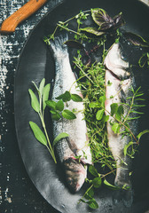 Cooking fish dinner. Flat-lay of raw uncooked sea bass fish with fresh green herbs on dark plate over rustic wooden painted background, top view. Helathy, clean eating, dieting food concept