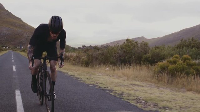 Tracking shot of a male cyclist sprinting on bicycle while climbing up a mountain road. Sportsman putting effort while climbing up the hill on a bicycle. 