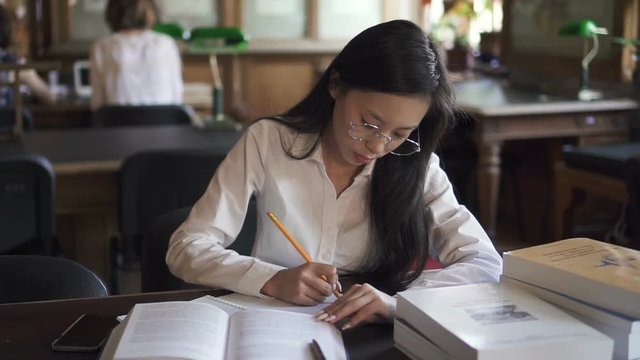 Female asian student lawyer in white shirt and round glasses studying in the library, taking notes. Indooors.