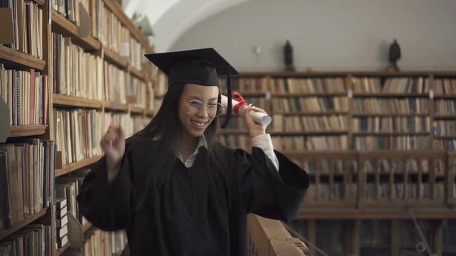 Female asian student lawyer in roung glasses and black gown holding scroll in her hand and enjoying her graduation in the library. Indoors. Portrait.