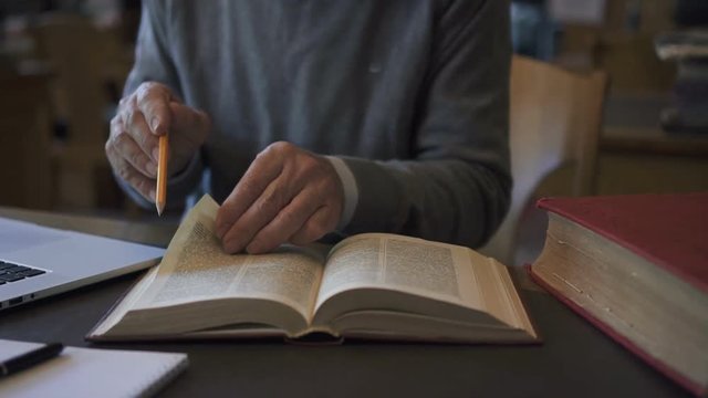 Elderly male biologist professor is examining book sitting at table in university library, intelligent man is looking, reading, pointing with pencil at desk with laptop during working day. Concept
