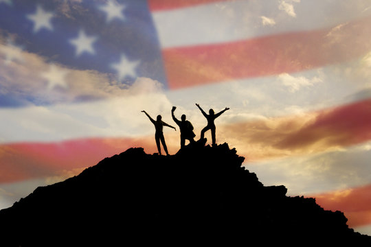 Silhouettes of happy three people on top of a mountain, but the background of the flag of the America and sky.