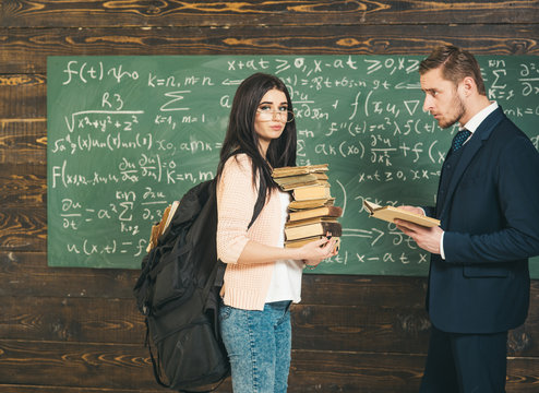 Brunette female student standing in front of green board with heap of books and huge backpack. Professor looking at beautiful girl in classroom
