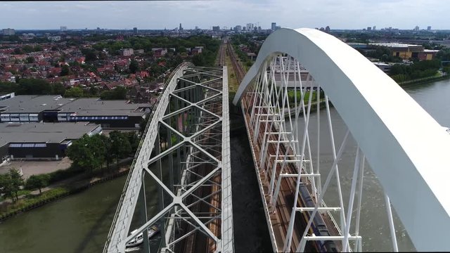 Aerial moving backwards over tied-arch railway bridge Utrecht city center skyline in background also showing train moving over tracks and recreational boats moving underneath the infrastructure 4k