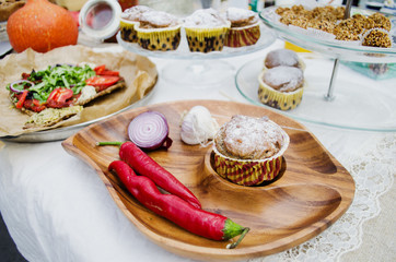 On a white tablecloth the muffin on a wooden plate with chilli pepper, onions and garlic. In the background, a plate with cupcakes and handmade chocolates and corn chips with arugula and tomatoes.