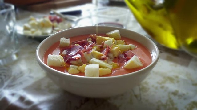 spreading olive oil on salmorejo cordobes, a typical spanish tomato soup similar to the gazpacho, topped with serrano ham, chopped boiled egg and croutons, on a set table