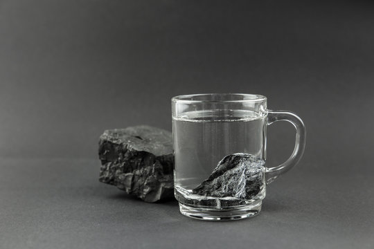 Water purification with black Shungite stones, clear drinking glass with stones on gray background. Removing toxins and organic matter from the water, black background.