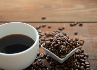 A cup of coffee with small white ceramic dish full of coffee beans on wooden background.