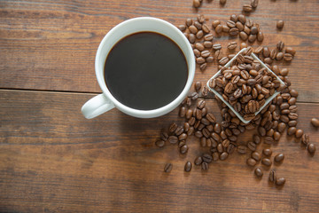 A cup of coffee with small white ceramic dish full of coffee beans on wooden background. Top view