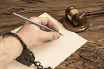 hand man handcuffed with a pencil, a blank sheet of paper, hammer judge on a wooden table...
