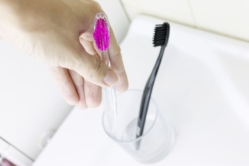 Cropped hand holding one of two toothbrushes of pink and black color on background of white washbowl and clear glass in bathroom.