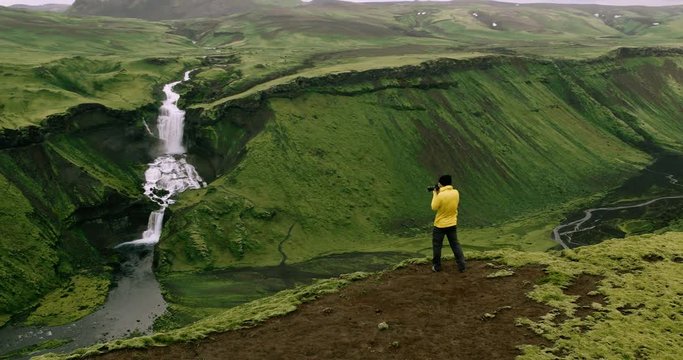 Aerial shot of adventure travel photographer standing on edge of cliff taking photos of waterfall mountain landscape
