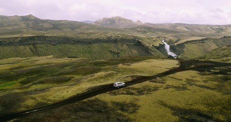 Aerial shot of 4x4 SUV car driving on top of mountain road with waterfall in background 