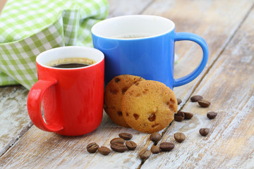 Two colorful mugs of black coffee on rustic wooden surface with biscuits and coffee beans scattered around
