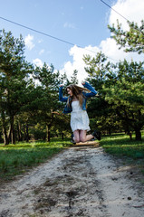 Cute and pretty young blond girl in white dress, jeans jacket and black boots jumping on the sand road in forest. Countryside girl have fun, she hop and turned her head sideways.