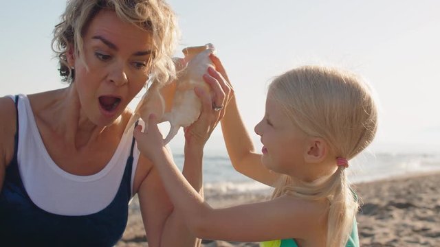 Grandmother and granddaughter sitting on beach playing with seashell