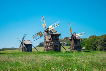 Antique old wooden windmills under blue sky on green field in front of forest. Sunny summer day. National Museum of Folk Architecture and Life of Ukraine in Pyrohiv. 