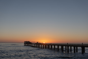 Fototapeta na wymiar Long pier above ocean waves at sunset with sun in center, Africa