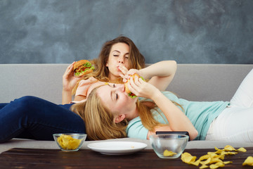 Overeating, sedentary lifestyle, fast-food, laziness, pleasure, delight, enjoyment, appetite, hunger. Two young lazy women lying at coach enjoy eating burgers and chips